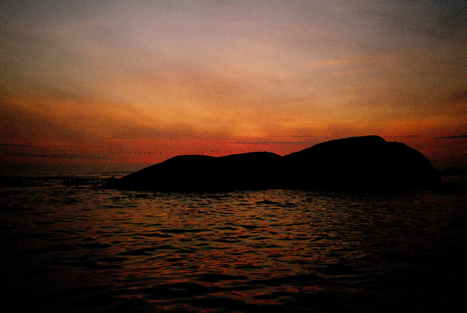 An orange sky above the ocean and the silhouette of a coastal rock.